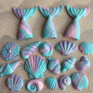 May include: A collection of mermaid tail and seashell decorations in teal, lavender, and pink. The assortment features various seashell designs and three mermaid tails, all with textured surfaces.
