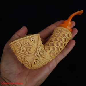 May include: A carved meerschaum pipe with an intricate floral design and a bright orange stem. The pipe is held in a hand.
