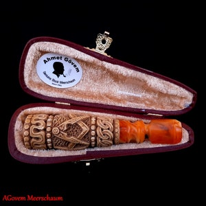Könnte beinhalten: Eine geschnitzte Meerschaumpfeife mit einem roten und bernsteinfarbenen Schaft, in einem burgunderfarbenen, samtenen Etui mit einem goldenen Verschluss. Das Etui trägt ein Etikett mit der Aufschrift "Ahmet Gövem Genuine Block Meerschaum Since 1963".