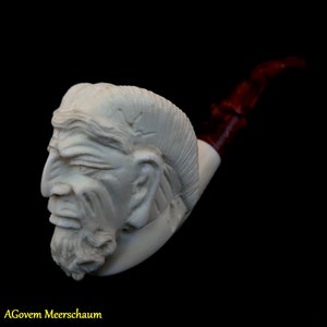 Puede incluir: Una pipa de espuma de mar blanca con una cara humana tallada. La pipa tiene una boquilla roja.