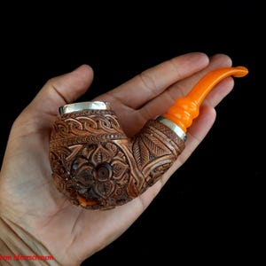 May include: A hand holds a brown wooden pipe with intricate carvings. The pipe has a silver band around the bowl and a bright orange acrylic stem.