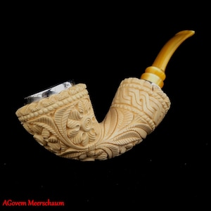 May include: A white meerschaum pipe with intricate floral carvings and a silver band. The pipe has a curved stem and a yellow mouthpiece.