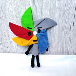 May include: A colourful plush pinwheel toy with a grey body and black legs. The blades are red, yellow, green, and blue. It has a white pom-pom in the centre and black eyes.