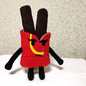 May include: A red plush toy shaped like a box of chocolate sticks. The toy has a yellow smiley face with black eyes and a black mustache. The toy has black arms and legs.