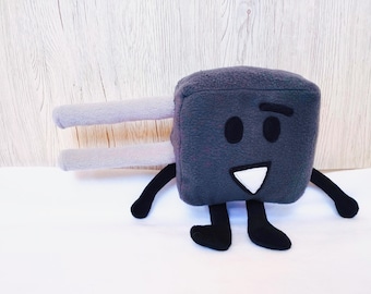May include: A soft toy in the shape of an electrical plug. The toy is dark grey with black eyes, a white triangle mouth, and black legs. It features two grey rectangular prongs extending from its side.