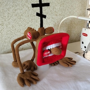 Pode incluir: Um peluche castanho com um corpo fino, braços longos e mãos grandes. A cabeça é uma estrutura retangular vermelha semelhante a um altifalante com uma boca larga, dentes brancos, uma língua vermelha e uma antena preta em forma de cruz.