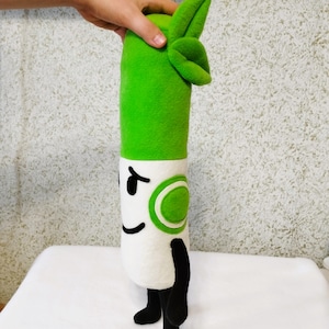 Pode incluir: Um brinquedo de pelúcia verde e branco em forma de pílula com uma folha verde no topo. O brinquedo tem um rosto sorridente com olhos pretos e um círculo verde de um lado.