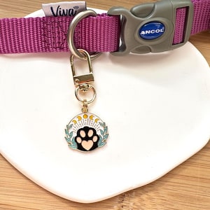 May include: Gold-tone dog tag featuring a black paw print and a white and blue floral design. The tag is attached to a purple and grey dog collar with the word "Viva" printed on the collar.
