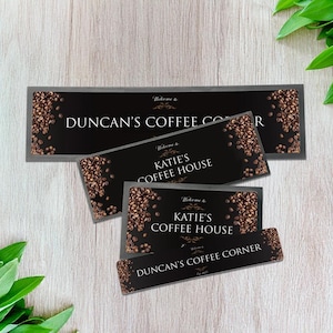Puede incluir: Varios letreros rectangulares negros con detalles de granos de café y texto. Los letreros dicen "Duncan's Coffee Corner" y "Katie's Coffee House" en escritura blanca. Sobre una superficie de madera clara.