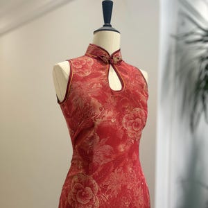 Puede incluir: Vestido rojo sin mangas de estilo chino, con cuello mandarín y estampado floral. El vestido se exhibe en un maniquí. Ribetes dorados acentúan el cuello y las sisas. Un detalle de ojo de cerradura está en el escote.