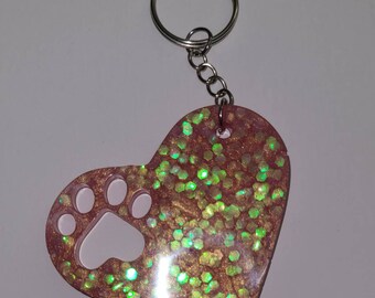 May include: Pink heart-shaped resin keychain with a glitter finish and a paw print cutout. The keychain has a silver metal ring.
