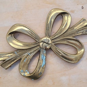 May include: A gold-toned metal bow-shaped trivet with a textured surface. The trivet is designed to look like a bow with a knot in the centre.