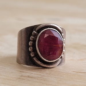 Puede incluir: Un anillo de plata con una gran piedra preciosa roja facetada engastada en un engaste de bisel. El anillo tiene una banda ancha con una superficie texturizada.