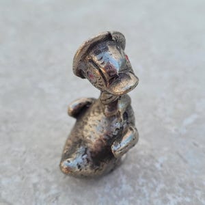 Puede incluir: Una figurita de pato de plata con ojos rojos y un sombrero. El pato está de pie sobre una superficie gris.