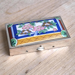 May include: A rectangular silver pillbox with a decorative ceramic lid. The lid features a floral design with pink and brown blossoms, green leaves, and a blue and white background. The box has a silver clasp.