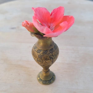 May include: A small, ornate brass vase with a floral design, holding a single pink flower.