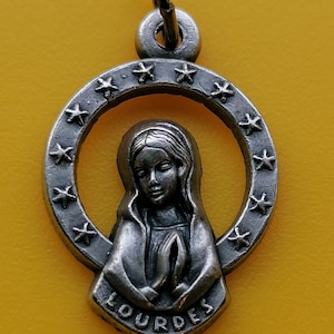May include: Silver-toned pendant featuring a depiction of the Virgin Mary with hands clasped in prayer. The pendant is encircled by a ring of stars and the word "LOURDES" at the bottom. The pendant hangs from a small loop.