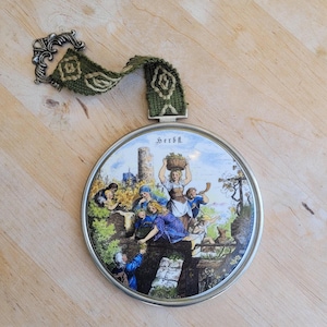 May include: A round, white ceramic wall hanging with a silver frame and a green and brown woven hanging strap. The ceramic features a painted scene of people harvesting grapes in a vineyard with a castle in the background. The text "Herbst" is written in black at the top of the ceramic.