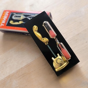 Op de afbeelding: Een zwarte doos met een gouden telefoonontvanger en een doorzichtige zandloper met rood zand. Op de doos staat "TIMER".