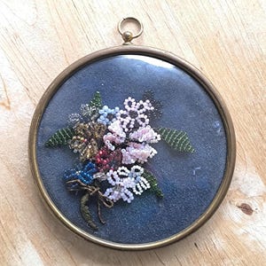 Puede incluir: Arreglo floral enmarcado hecho de cuentas de colores. El arreglo presenta flores y hojas de cuentas rosas, blancas, azules y verdes, sobre un fondo azul oscuro. El marco es redondo con un acabado de latón y un pequeño lazo en la parte superior.