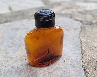 May include: A small, amber-coloured glass bottle with a black screw-top lid. The bottle is slightly rounded and has a slight curve to the bottom.
