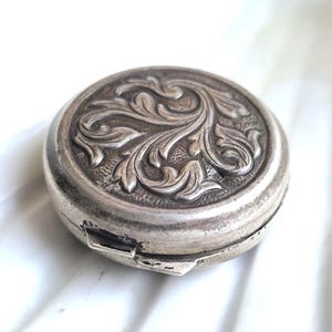 Op de afbeelding: Een kleine, ronde, zilverkleurige compact met een sierlijk, verhoogd bloemmotief op het deksel. De compact heeft een scharnierende sluiting en een licht aangetaste uitstraling, wat op leeftijd duidt. Het ontwerp toont wervelende bladeren en ranken.