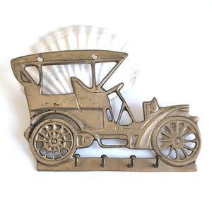 May include: A vintage-style brass key holder shaped like an antique car. The key holder features a detailed design of the car's body, wheels, and roof, with three hooks below for keys. The brass has a slightly aged appearance.