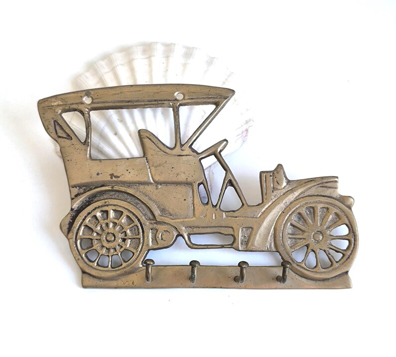 May include: A vintage-style brass key holder shaped like an antique car. The key holder features a detailed design of the car's body, wheels, and roof, with three hooks below for keys. The brass has a slightly aged appearance.