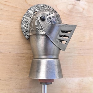 Peut inclure: Un bouchon de bouteille en métal argenté surmonté d'un casque de chevalier détaillé. Le casque présente une crête gaufrée ornée, une visière articulée avec trois fentes horizontales et une base lisse évasée fixée à une tige métallique fine.