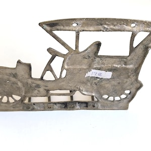 May include: A silver-colored metal wall hanging in the shape of a vintage car. The car has a detailed design with visible wheels, a driver's seat, and a roof. The piece has a distressed finish and mounting holes.