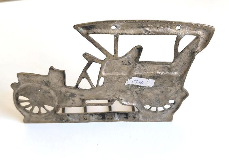 May include: A silver-colored metal wall hanging in the shape of a vintage car. The car has a detailed design with visible wheels, a driver's seat, and a roof. The piece has a distressed finish and mounting holes.