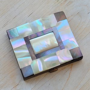 May include: A square, vintage compact with a mother-of-pearl mosaic design. The iridescent tiles are arranged in a geometric pattern, with a rectangular centerpiece. The compact has a metal frame and a clasp closure.