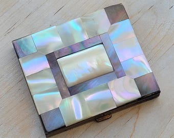 May include: A square, vintage compact with a mother-of-pearl mosaic design. The iridescent tiles are arranged in a geometric pattern, with a rectangular centerpiece. The compact has a metal frame and a clasp closure.