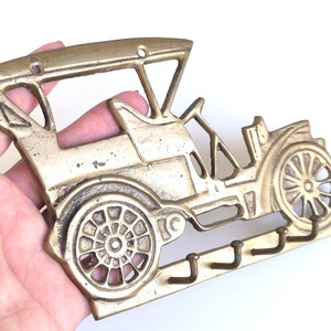 May include: A brass-colored key holder shaped like a vintage car. The key holder features a detailed car design with wheels, a roof, and three hooks for keys. The metal has a slightly aged appearance.