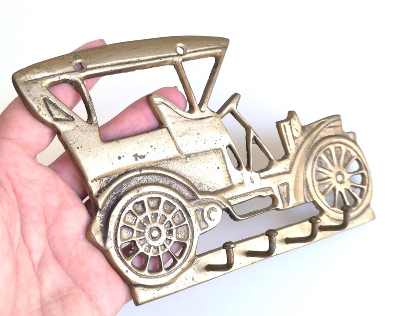 May include: A brass-colored key holder shaped like a vintage car. The key holder features a detailed car design with wheels, a roof, and three hooks for keys. The metal has a slightly aged appearance.