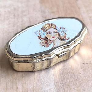Könnte beinhalten: Eine ovale, vintage Pillendose aus goldfarbenem Metall. Der Deckel zeigt ein gemaltes Porträt einer Frau mit Blumen im Haar. Die Dose hat einen gewellten Rand und einen Schnappverschluss, der auf einer Holzoberfläche liegt.