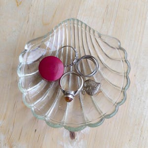 Pode incluir: Um prato de vidro transparente em forma de concha com uma coleção de anéis e um objeto redondo de cor magenta. Os anéis são prateados e incluem um anel com uma pedra preciosa, um anel com um design texturizado e um conjunto de anéis finos. Também está presente um pequeno pingente prateado em forma de folha.