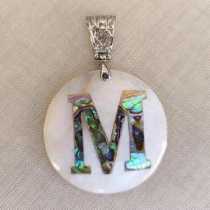 Op de afbeelding: Een witte parelmoer hanger met een zilveren beugel en een grote letter M gemaakt van iriserende abalone schelp.