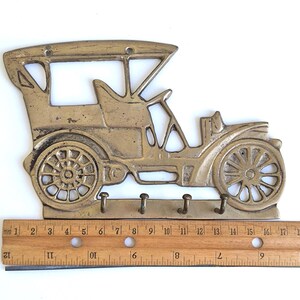 May include: A brass-colored key holder shaped like a vintage car. The key holder has three hooks below the car's body. The car design includes wheels, a steering wheel, and a roof. The key holder measures approximately 19 inches in length.