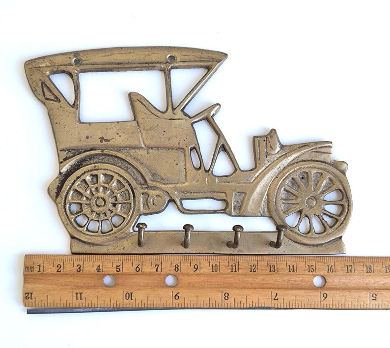 May include: A brass-colored key holder shaped like a vintage car. The key holder has three hooks below the car's body. The car design includes wheels, a steering wheel, and a roof. The key holder measures approximately 19 inches in length.