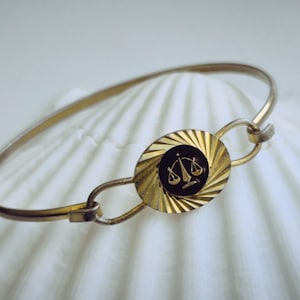 May include: A gold bracelet with a black and gold circular charm featuring a stylized scale design.