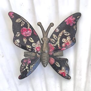 May include: A decorative butterfly pin with a dark background and pink and gold floral accents. The butterfly has a bronze-colored body and antenna. The wings are detailed with a floral pattern.