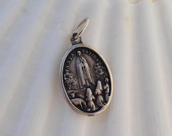 Può includere: Pendente ovale in argento con una raffigurazione della Vergine Maria e tre bambini. Il pendente ha le parole "Nossa Senhora de Fatima" incise attorno al bordo.