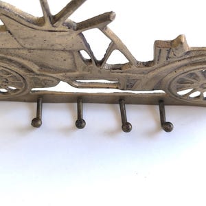 May include: A vintage-style key holder shaped like a classic car. The metal key rack is a muted gold color and features four hooks below the car's chassis. The car's design includes detailed wheels and a roof.