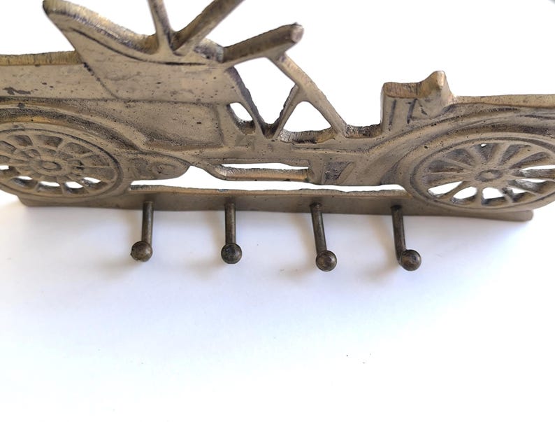 May include: A vintage-style key holder shaped like a classic car. The metal key rack is a muted gold color and features four hooks below the car's chassis. The car's design includes detailed wheels and a roof.