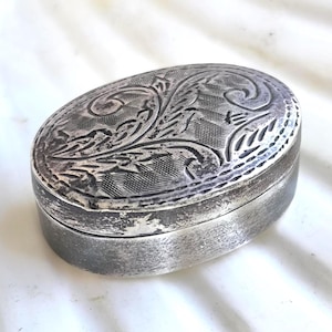 Può includere: Un'antica scatola ovale in argento. Il coperchio presenta un ornato disegno floreale inciso. La scatola ha un coperchio incernierato e una finitura leggermente ossidata, che le conferisce un aspetto vintage. Probabilmente usata per riporre piccoli oggetti.
