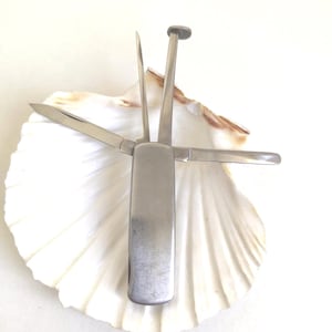 May include: A silver multi-tool with a knife blade, reamer, tamper, and pick. The tool is open, displaying its various components. The tool is resting on a white seashell.