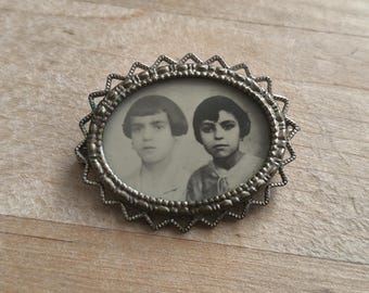 May include: A silver oval locket with a scalloped edge, featuring a black and white photo of two children. The locket is a vintage style piece of jewellery.