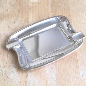 Könnte beinhalten: Ein rechteckiger Aschenbecher aus silberfarbenem Metall mit einer polierten, reflektierenden Oberfläche. Die Schale verfügt über einen glatten, leicht geschwungenen Rand und zwei integrierte Einkerbungen an gegenüberliegenden Seiten.