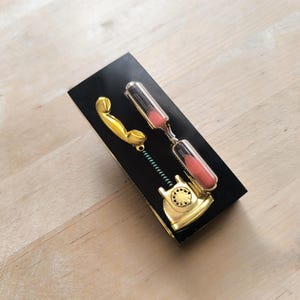 May include: A gold-colored phone receiver with a black hourglass attached to it. The hourglass is filled with pink sand. The phone receiver and hourglass are on a black background.